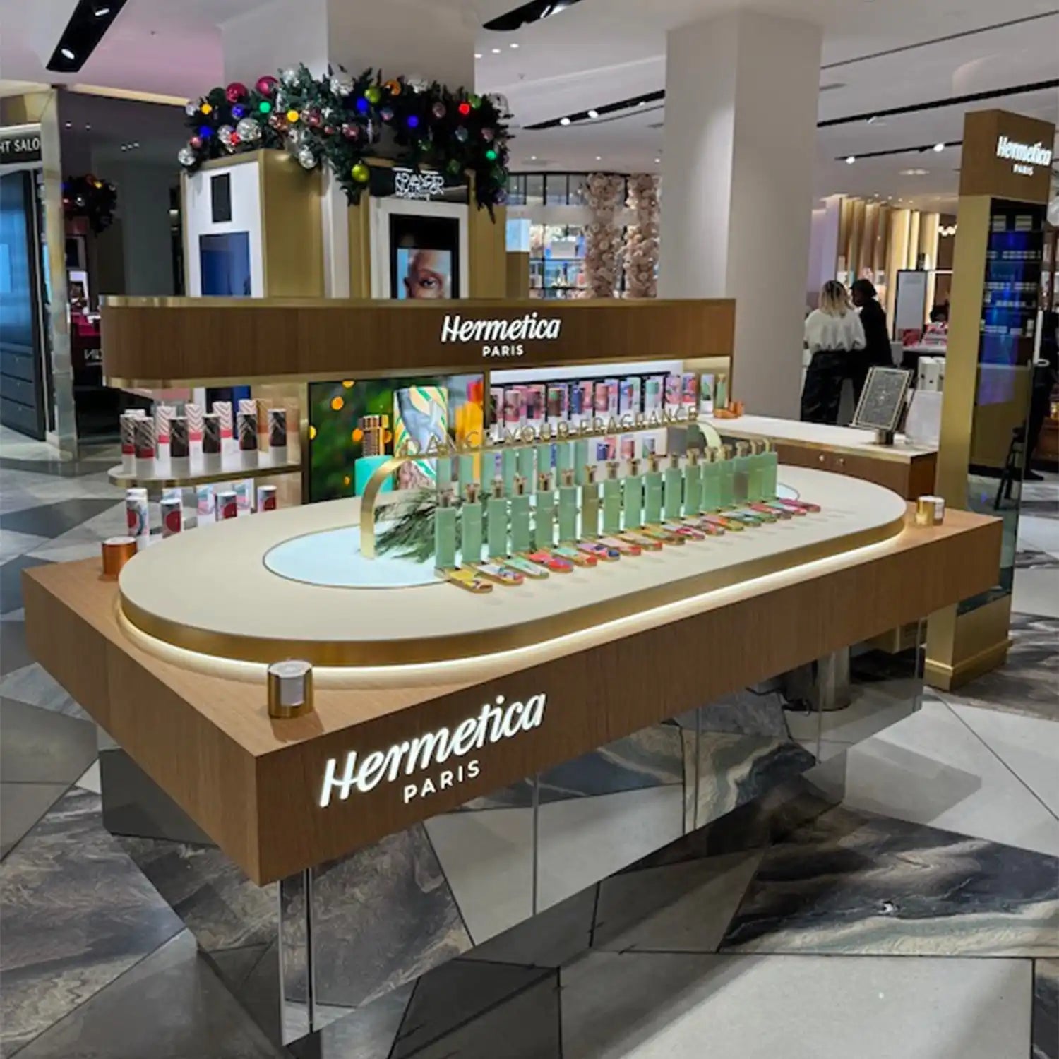 A retail display kiosk for Hermetica perfumes featuring an oval-shaped counter with product bottles arranged in rows.